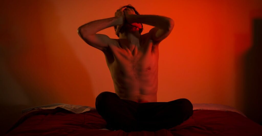 A topless man showing emotional distress sitting on a bed in a dimly lit room.